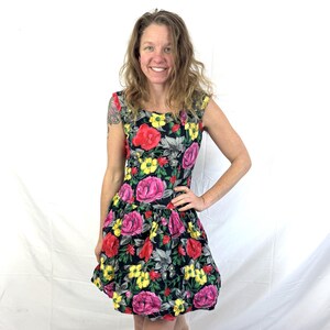 May include: A sleeveless floral dress featuring a black background with large pink, red, and yellow flowers. The dress has a square neckline and a flared skirt. The model is barefoot.