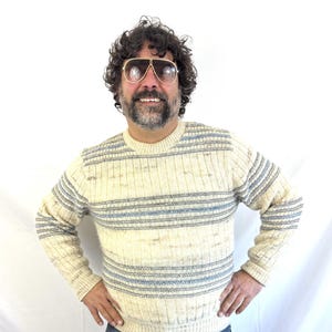 以下が含まれることがあります： 水平な青いストライプが入ったクリーム色のセーター。長袖ニットはリブ編みのテクスチャとクルーネックです。セーターはブルージーンズと合わせています。その人はアビエーターサングラスをかけています。