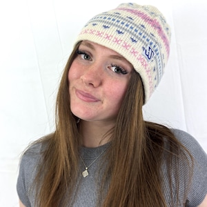 May include: A cream-coloured knitted beanie hat with a geometric pattern in blue and pink. The hat has a small blue logo on the side. The beanie is a winter accessory.