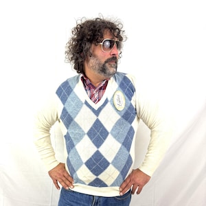 May include: Cream-colored argyle sweater vest with blue and white diamond pattern. The sweater is worn over a red and black plaid shirt and cream-colored long-sleeve shirt. The person is wearing blue jeans and sunglasses.