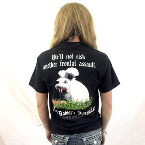 Peut inclure: Un t-shirt noir à manches courtes avec un motif de lapin blanc aux yeux rouges et aux dents acérées dans de l'herbe verte. Le texte indique : We'll not risk another frontal assault. That Rabbit's Dynamite! Porté avec un jean en denim bleu.