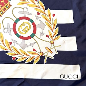 May include: A navy blue and white striped silk scarf featuring a central nautical emblem with a red and gold crown, a life preserver with a gold interlocking logo, a silver anchor, and yellow laurel leaves. The word GUCCI is printed in black.
