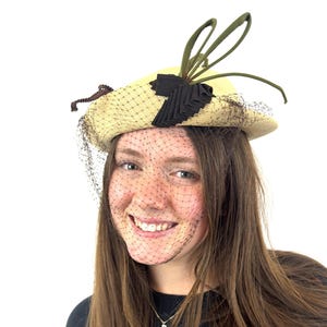 May include: A woman wearing a straw hat with a black ribbon and a green feather. The hat has a black veil. She is smiling and looking at the camera.