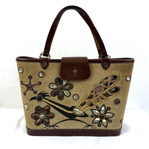 May include: A tan tote bag with a brown leather handle and accents. The bag features a roadrunner design with floral and jewel embellishments. The bag has a brown leather flap closure.