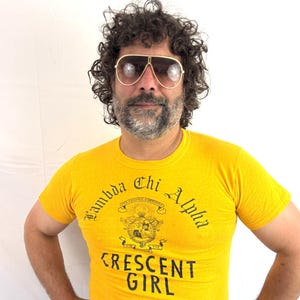 May include: A bright yellow short-sleeved t-shirt featuring the black text Lambda Chi Alpha and Crescent Girl, along with a central crest graphic. The shirt is worn with dark blue denim jeans and large gold-rimmed aviator sunglasses.