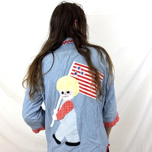 May include: A light blue button-down shirt featuring a large fabric appliqué on the back of a blonde child holding a red and white striped flag with blue stars. The shirt has red and white polka dot cuffs and a red scalloped trim along the hem.