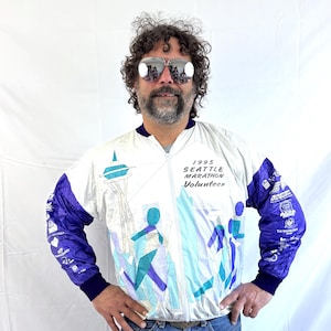 Puede incluir: Una chaqueta cortavientos blanca con un diseño morado y azul. La chaqueta tiene una cremallera y un cuello alto. La chaqueta presenta un gráfico del Space Needle y el texto "1995 Seattle Marathon Volunteer".