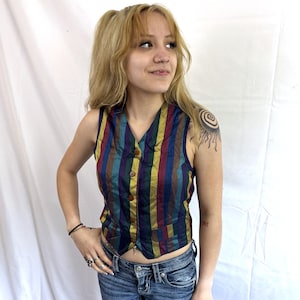 May include: A sleeveless vest featuring vertical stripes in shades of dark blue, burgundy, and gold. The garment has a V-neckline, a front button closure with four brown buttons, and two small welt pockets at the waist. The fabric has a slight sheen.