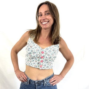 Op de afbeelding: Een mouwloze crop top met een witte basis en een klein bloemenpatroon in blauwe en roze tinten. Het kledingstuk heeft een V-hals en een knoopsluiting aan de voorzijde met vier ronde, effen roze knopen verticaal in het midden geplaatst.