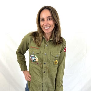 May include: An olive green long-sleeve button-down shirt featuring two chest pockets, a collar, and various embroidered patches on the front and sleeve. The shirt includes a red patch with the number 2 and a circular patch with a landscape graphic.