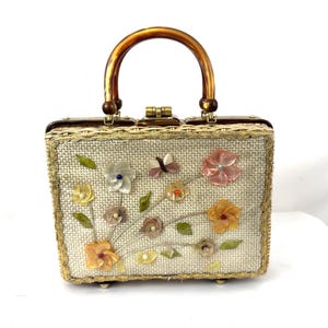 Può includere: Una borsetta vintage a forma di scatola con corpo in paglia intrecciata e manico marrone. La parte anteriore della borsa è decorata con fiori di stoffa colorati e una farfalla. La borsa ha un bordo dorato e una chiusura a scatto.