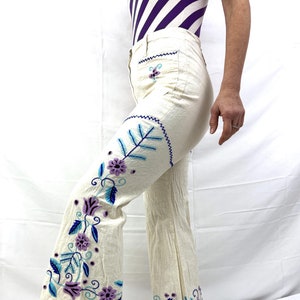 Puede incluir: Pantalón blanco acampanado con diseños florales bordados en tonos de morado, azul y verde.