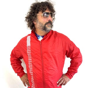 May include: A bright red zip-up jacket with white stripes and the word "Cools" printed repeatedly down the left side. The jacket is lightweight and has a classic, vintage style. The model is wearing blue jeans.