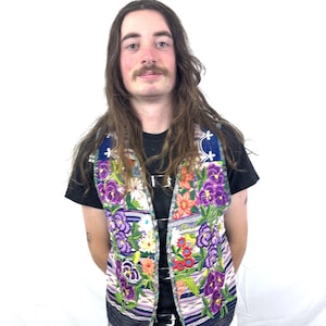May include: A colorful, embroidered vest with a floral pattern. The vest features a variety of flowers in shades of purple, orange, and white against a striped background. The vest is worn over a black t-shirt and dark blue jeans.