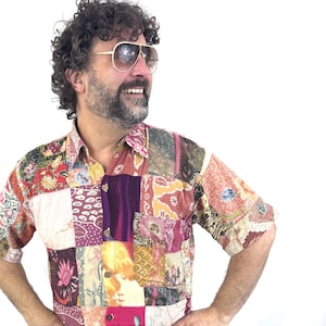 Può includere: Una camicia patchwork con maniche corte. La camicia presenta una varietà di motivi in tessuto nei toni del rosso, rosa, viola e crema. La camicia è indossata con jeans blu.