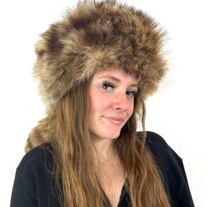 Può includere: Un cappello in finta pelliccia marrone con corona arrotondata e paraorecchie. Il cappello è realizzato in un materiale morbido e strutturato e ha un aspetto naturale e soffice. Il cappello è progettato per tenere al caldo la testa e le orecchie. È un accessorio invernale.