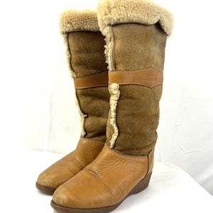 May include: A pair of tall, brown leather boots with a sheepskin lining and trim. The boots have a light brown leather foot and a darker brown suede upper, with a strap detail. The boots are designed for cold weather.
