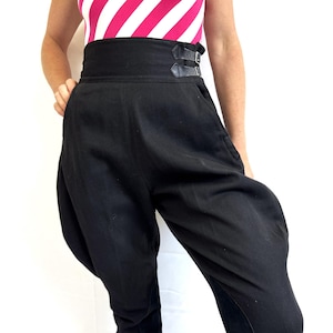 May include: Black high-waisted pants with a wide waistband and two leather buckle closures. The pants have a unique, voluminous design with button details at the ankles. The model is wearing a pink and white striped top.