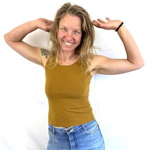 May include: Mustard-colored tank top and blue denim jeans. The tank top has a round neckline and a fitted silhouette. The jeans are a light wash with a button closure. A black bracelet is visible on the left wrist.