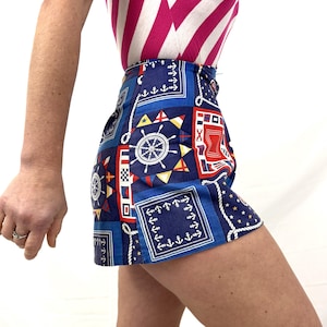 May include: Blue nautical-themed shorts with white rope trim and a repeating pattern of anchors, ship wheels, and geometric shapes. The shorts are high-waisted and feature a button closure. The shorts are paired with a pink and white striped top.