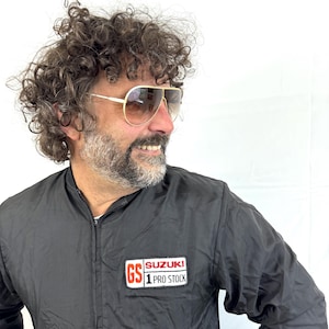 May include: Gold-framed aviator sunglasses with gradient lenses. The person is wearing a black nylon jacket with a "GS SUZUKI 1 PRO STOCK" patch on the chest. The jacket has a zipper closure.