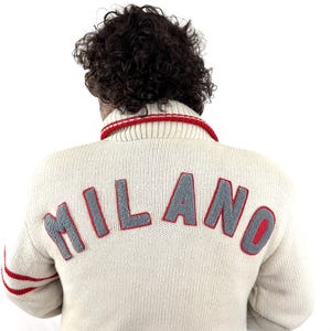May include: Cream-colored sweater with the word "MILANO" in gray letters outlined in red across the back. The sweater has a red and white striped collar and red stripes on the sleeves. A classic, vintage-inspired design.