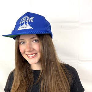May include: A blue and white baseball cap with the IBM logo and a ship design. The logo is in white and the ship is in gray. The cap has a green brim.