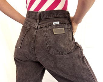 May include: High-waisted dark brown denim jeans featuring a Wrangler brand patch on the back pocket, a leather-like rectangular patch above the pocket, belt loops, and a classic five-pocket construction with visible stitching throughout the garment.