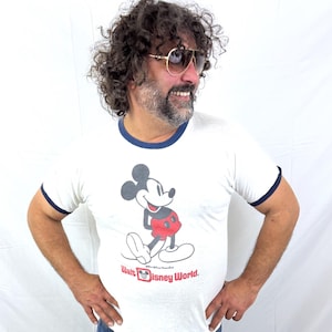 May include: White ringer t-shirt featuring a graphic of Mickey Mouse with red shorts and the text "Walt Disney World." The shirt has navy blue trim around the collar and sleeves. The person is wearing blue jeans and gold-rimmed sunglasses.