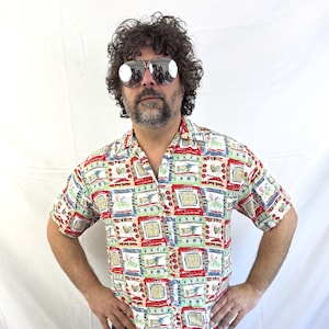 May include: Short-sleeved button-down shirt with a colorful geometric pattern in red, green, blue, and white. The shirt has a classic collar and is paired with blue jeans. The shirt's design features repeating rectangular shapes with various illustrations.