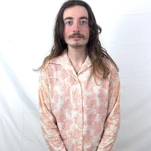 Può includere: Camicia a maniche lunghe con motivo floreale pesca e bianco. La camicia ha un colletto classico ed è abbottonata. Il tessuto sembra leggero e viene indossato con pantaloni marroni.