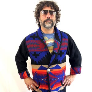May include: A black wool jacket with a vibrant geometric pattern in red, blue, orange, and beige. The jacket features a shawl collar, a single silver button, and red and orange cuffs. The jacket is worn over a blue t-shirt and blue jeans.