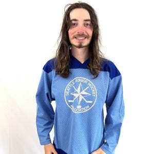 Puede incluir: Camiseta de hockey azul claro con un logotipo blanco que dice "Seattle Junior Hockey Association". La camiseta tiene cuello en V y detalles azules en los hombros. Es de manga larga y está hecha de un material perforado y transpirable.