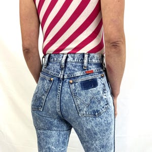 Puede incluir: Vaqueros de mezclilla con lavado &aacute;cido, de cintura alta y pernera recta. Los vaqueros tienen un efecto marmoleado azul y blanco, con un parche azul oscuro en el bolsillo trasero. La persona lleva una camiseta sin mangas a rayas rosas y blancas.