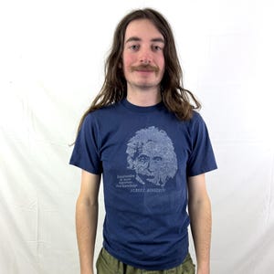 Pode incluir: Camiseta azul marinho com um gráfico azul claro de Albert Einstein e a citação "A imaginação é mais importante que o conhecimento". O texto "ALBERT EINSTEIN" está abaixo. A pessoa está usando calças cargo verde-oliva.