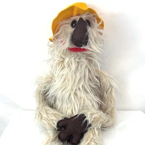 May include: A handmade, whimsical plush doll with a shaggy, cream-colored body. It features a yellow felt hat, a brown nose, and a red mouth. The doll's arms are crossed, and it has brown felt hands. The doll is a unique, handcrafted item.