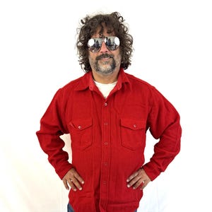 May include: A long-sleeved, solid red button-down shirt featuring a pointed collar, two chest pockets with buttoned flaps, and a front button closure. The shirt is worn over a white undershirt and paired with blue denim jeans.