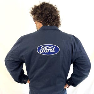May include: A navy blue work jacket featuring a classic collar, long sleeves with buttoned cuffs, and a prominent oval Ford logo patch in blue and white centered on the back. The jacket is constructed from a durable fabric and includes a straight hem.