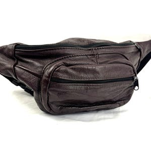 May include: A dark brown bum bag with a textured surface, featuring a main zipped compartment and a smaller front zipped pocket. The bag includes a black adjustable strap with a plastic buckle closure, designed for waist or crossbody wear.