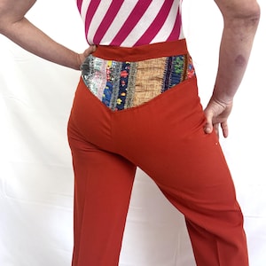 May include: Rust-colored pants with a decorative patchwork waistband. The pants have a flared leg and a high waist. The top is a pink and white striped shirt. The outfit is a vintage style.
