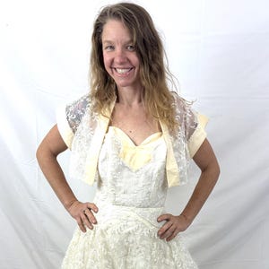 Puede incluir: Vestido de novia vintage de color marfil con una superposici&oacute;n de encaje y un escote coraz&oacute;n. El vestido presenta una falda amplia y se combina con un bolero de encaje a juego con ribetes amarillo p&aacute;lido. Un conjunto nupcial cl&aacute;sico.