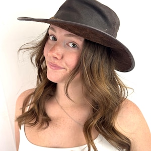 Puede incluir: Un sombrero fedora de color marr&oacute;n oscuro con ala ancha y acabado texturizado. El sombrero presenta una copa estructurada y un aspecto ligeramente desgastado a lo largo del borde del ala, mostrando un color marr&oacute;n intenso en toda la pieza.