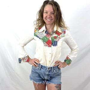 May include: Cream-colored button-down shirt with patchwork detailing on the collar and yoke. The patchwork features red, green, and blue floral and geometric patterns. The long sleeves have matching patchwork cuffs. Worn with denim shorts.