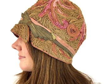 Puede incluir: Un sombrero cloche vintage con intrincados bordados en tonos dorados, rosas y melocotón. El sombrero presenta una banda decorativa con un acento de hoja verde y una cinta rosa. Un accesorio elegante.