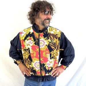 May include: A vintage black and gold zip-up jacket with a vibrant baroque-style pattern in red, gold, and white. The jacket has black sleeves and a black collar. The cuffs have a red and gold design. The jacket is worn with blue jeans.