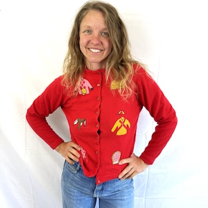 May include: A red cardigan sweater with various embroidered patches. The patches include a pink jacket, a brown saddle, a yellow riding outfit, and a pink patch with text. The sweater has long sleeves and a button-up front.