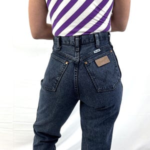 Puede incluir: Vaqueros Wrangler de mezclilla oscura con cintura alta y parche de cuero. Los vaqueros tienen remaches color lat&oacute;n y una peque&ntilde;a etiqueta Wrangler. Se ve una camiseta sin mangas con rayas diagonales moradas y blancas.