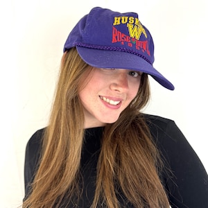 Può includere: Un cappellino da baseball viola con la scritta "HUSKIES ROSE BOWL 1991" ricamata in giallo, rosso e verde. Il cappellino ha un dettaglio a corda intorno all'orlo.