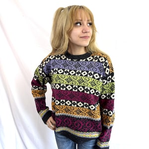 Può includere: Un maglione a maniche lunghe con girocollo caratterizzato da un motivo orizzontale di forme geometriche e disegni a spirale nei toni del viola, verde lime, magenta e arancione, su sfondo grigio scuro con polsini e scollo a coste.