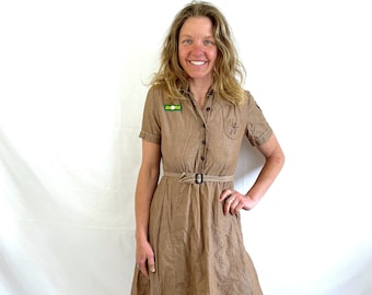 May include: A tan short-sleeved dress with a button-up front, a collared neckline, and a matching belt. The dress features a small patch on the chest and a decorative emblem. The dress is knee-length and has a slightly flared skirt.
